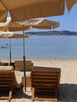 Terrasse extérieure avec parasols et chaises longues sur le sable, vue sur la mer calme et un îlot en arrière-plan.