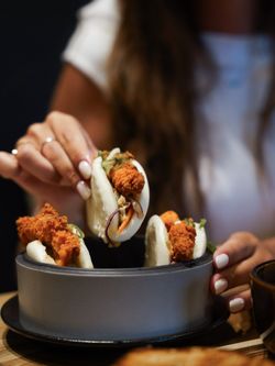 Trois baos garnis de poulet frit et légumes, servis dans un bol gris, avec une personne tenant un bao.