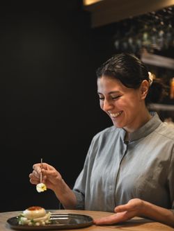 Chef souriante en veste grise préparant un plat raffiné dans un restaurant asiatique moderne.