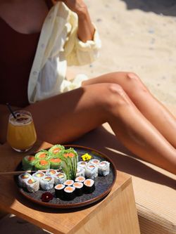 Sushi variés et rouleaux de printemps sur assiette noire, près d'une personne assise sur une terrasse ensoleillée.